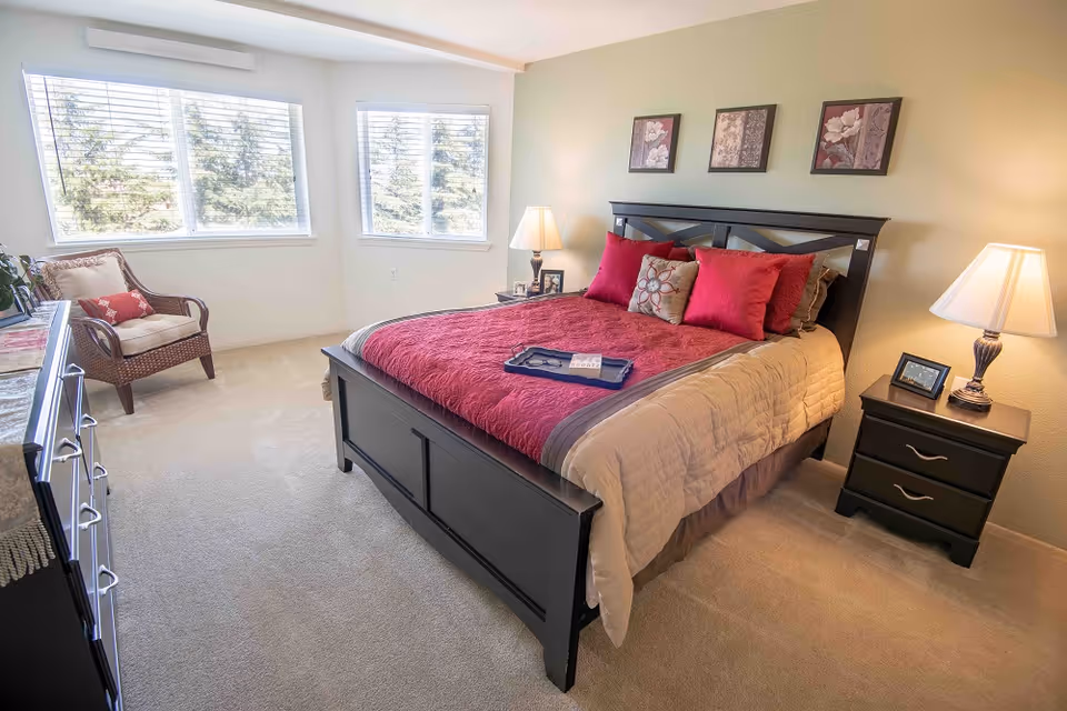 A bright and cozy bedroom with a large bed featuring a red and beige comforter and multiple pillows. The room has beige carpet, two bedside tables with lamps, a wicker chair with cushions near two large windows, and three framed floral artworks on the wall above the bed.