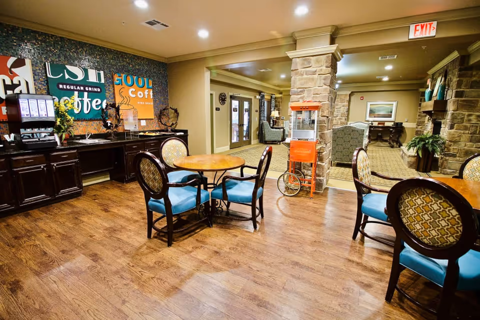 Communal dining and coffee area with round tables, turquoise-upholstered chairs, a coffee station against the wall and a popcorn cart near stone columns.