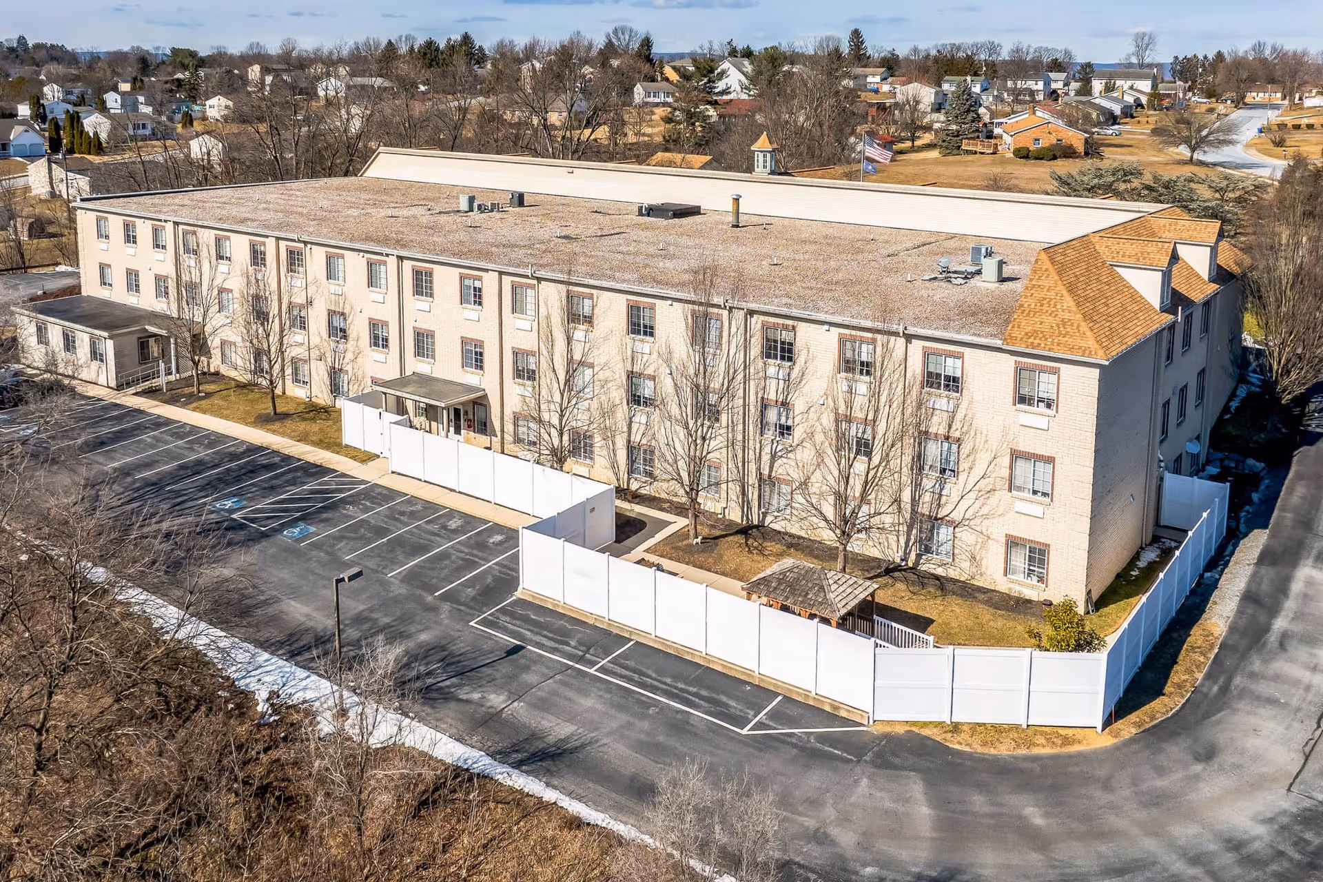 Aerial view of a large, three-story senior living facility building with a flat roof and beige exterior. The building is surrounded by a white fence enclosing a small yard area with a gazebo. There is a parking lot with marked spaces, including handicapped spots, adjacent to the building. The surrounding area includes residential houses and trees with no leaves, indicating a winter or early spring season.