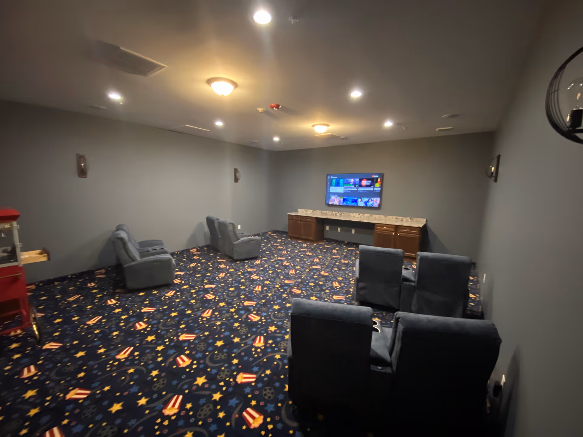 Small media room with upholstered recliner seats facing a wall-mounted TV and a colorful patterned carpet.