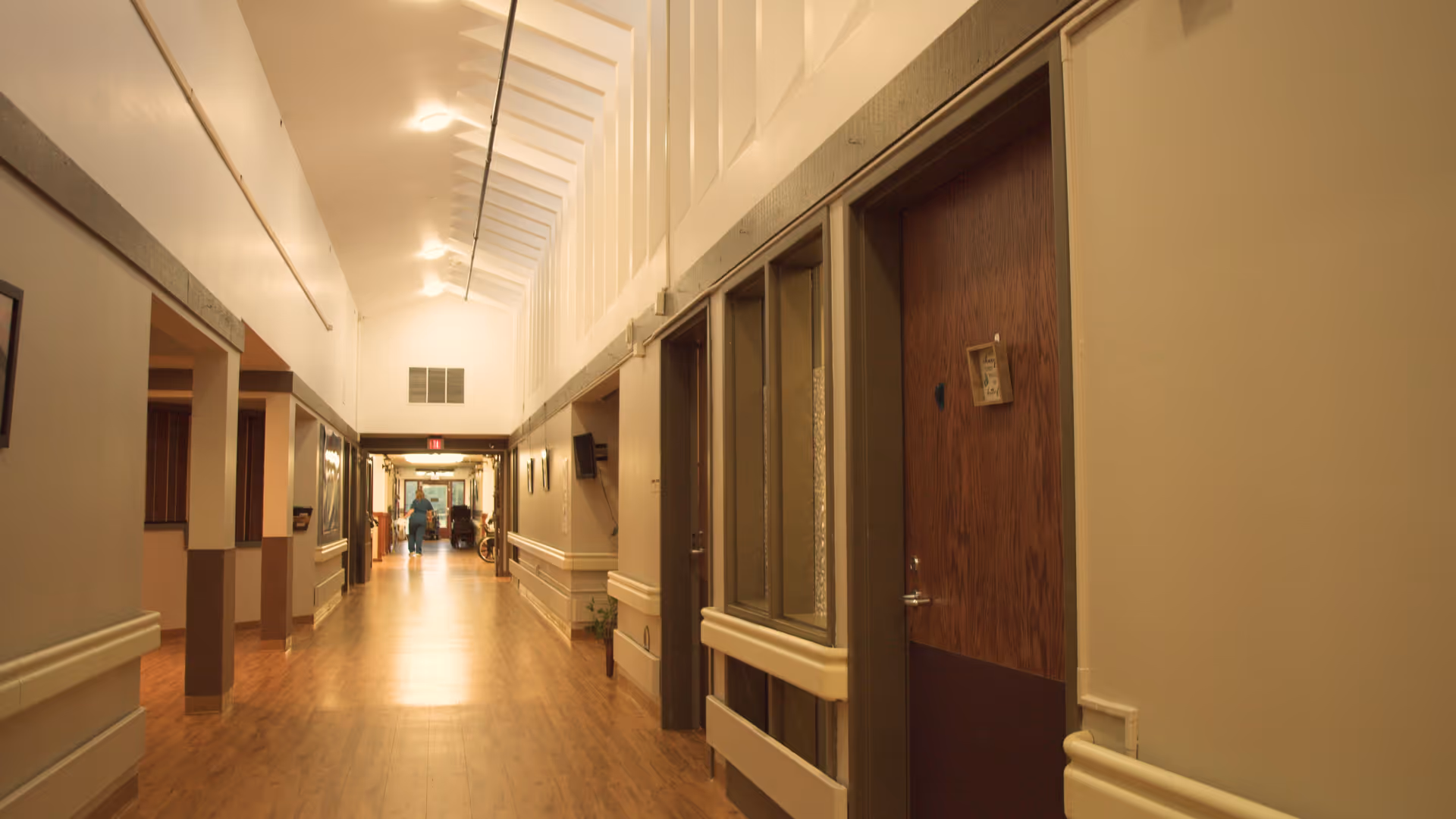 A well-lit hallway in a senior living facility with wooden flooring, beige walls, and several doors along the right side. The ceiling has a series of skylights allowing natural light to brighten the corridor. At the far end, a person in a wheelchair and another person are visible.
