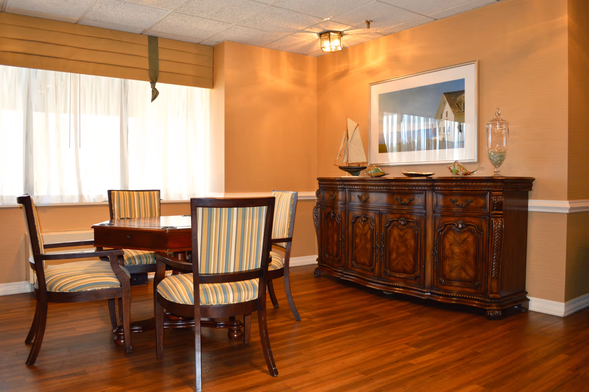 A cozy dining area with a wooden table surrounded by four striped upholstered chairs. The room features warm beige walls, a large window with sheer white curtains, and a wooden sideboard with decorative items including a model sailboat and a framed picture above it. The floor is wooden, and the lighting is soft and warm.