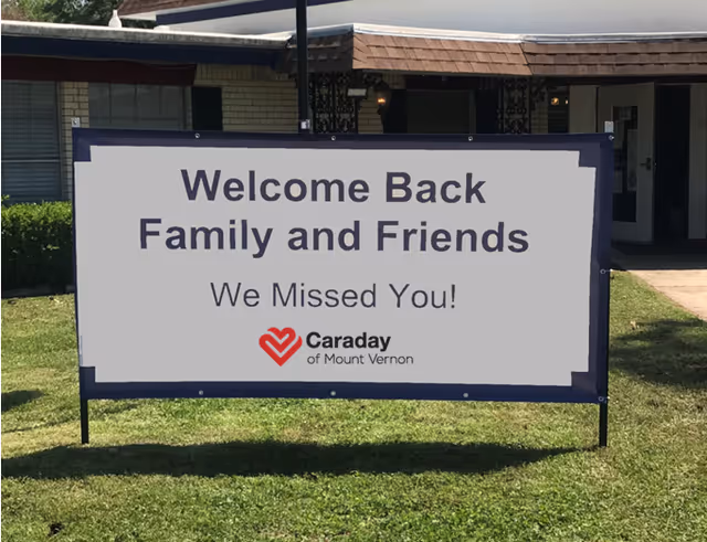 A large outdoor sign on a lawn in front of a building reads 'Welcome Back Family and Friends We Missed You!' with the Caraday of Mount Vernon logo below the text.