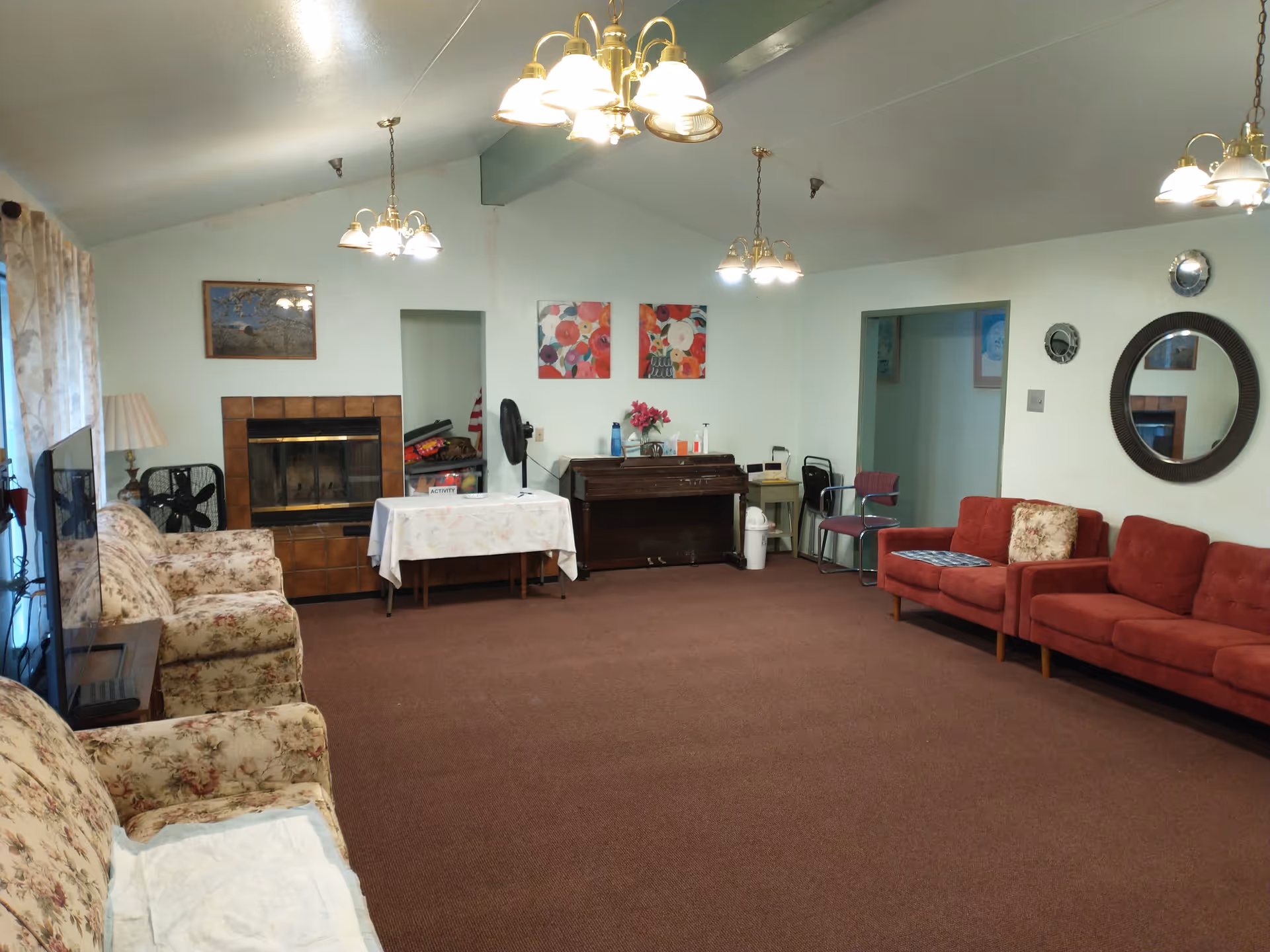 A spacious living room with floral patterned armchairs on the left and red sofas on the right. The room has a brown carpet, a fireplace with a tiled surround on the far wall, and a piano against the back wall. There are several light fixtures hanging from the ceiling, a round mirror on the right wall, and various decorative paintings and flowers. A table covered with a floral tablecloth is positioned near the fireplace.