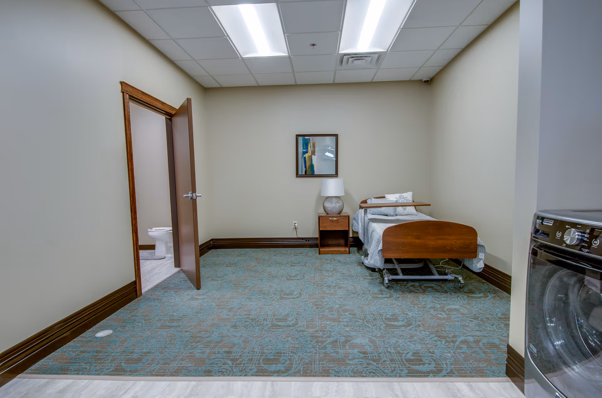 A small room with a single hospital-style bed with wooden headboard and footboard, a bedside table with a lamp, and a framed abstract painting on the wall. The room has beige walls, a patterned blue carpet, and a door leading to a bathroom with a visible toilet. A washing machine or dryer is partially visible on the right side.