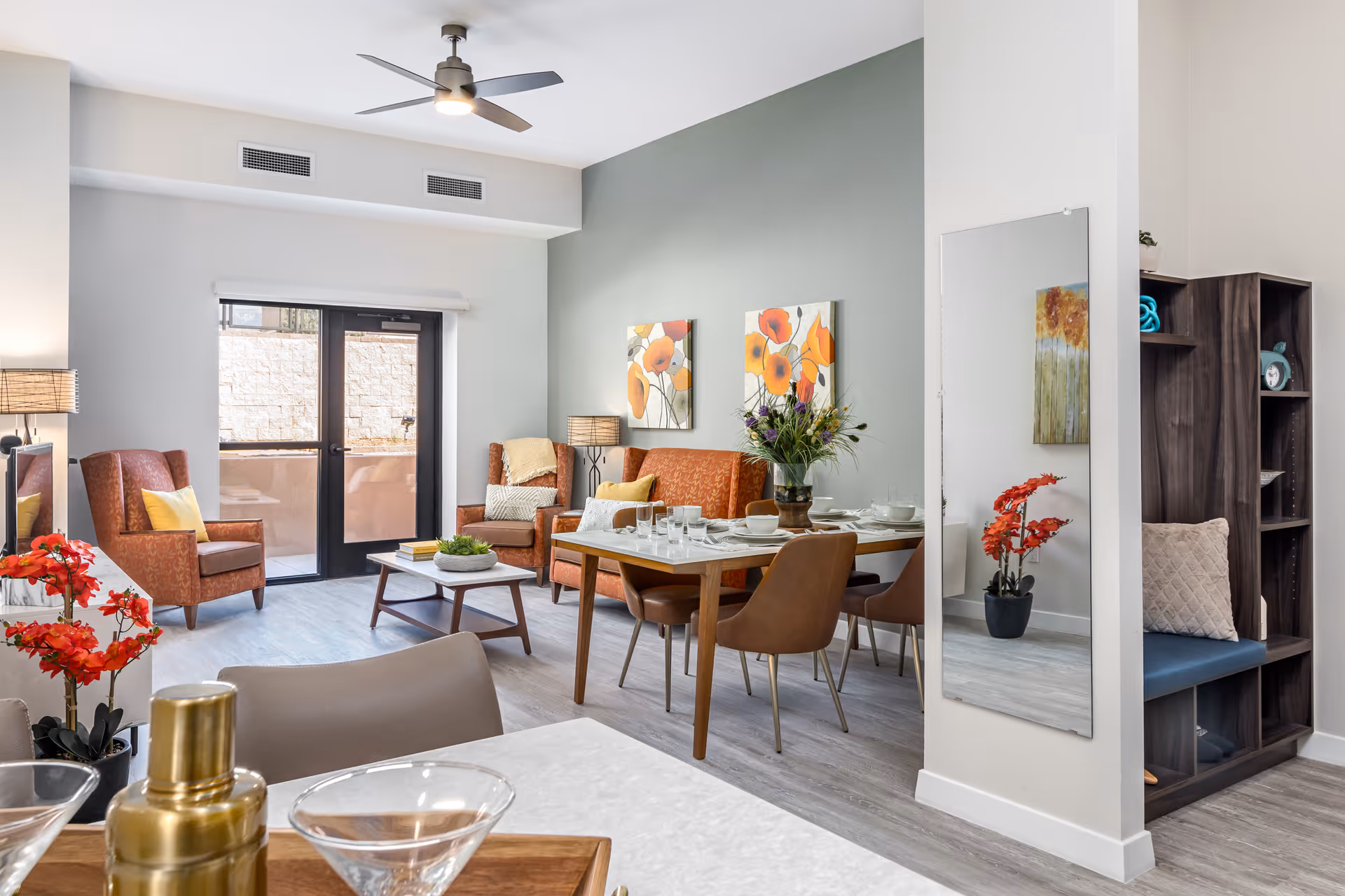 A bright and modern living and dining area in a senior living facility. The space features two brown armchairs with yellow pillows, a brown sofa, a wooden coffee table, and a dining table set with plates and glasses. There are colorful floral paintings on the wall, a ceiling fan, and a large window with a door leading outside. A tall mirror and a shelving unit with cushions and decorative items are also visible.