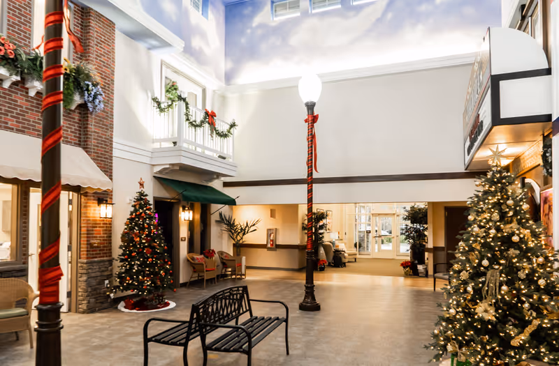 Indoor common area decorated for Christmas with two decorated Christmas trees, a black metal bench, a lamppost wrapped with red ribbon, and garlands with red bows hanging on the balcony railing. The area has a high ceiling painted with clouds and a bright, welcoming atmosphere.
