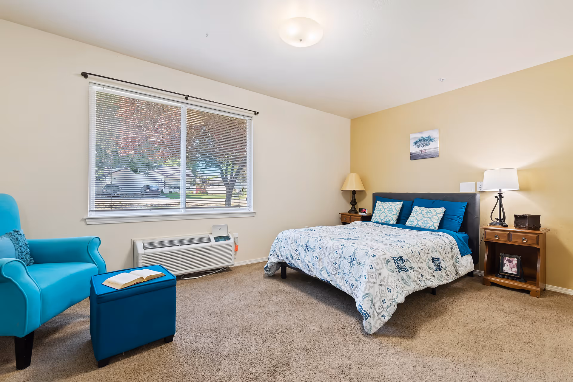 A cozy bedroom in Autumn Wind Assisted Living featuring a bed with blue and white patterned bedding and pillows, two wooden nightstands with lamps on either side, a turquoise armchair with a matching ottoman holding an open book, a large window with blinds showing trees and parked cars outside, and beige carpeted floor.