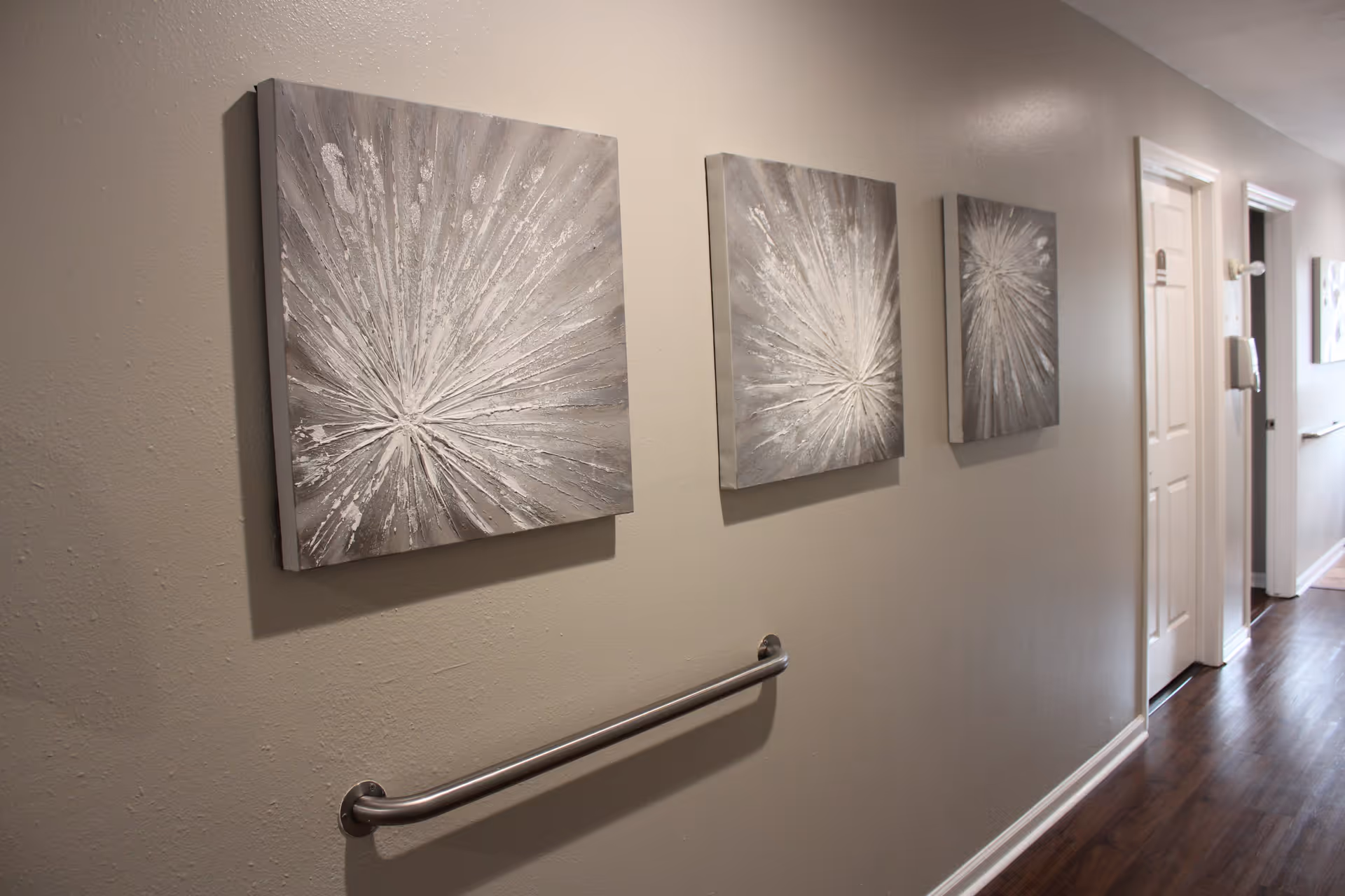 A hallway in an assisted living facility with three abstract silver and gray paintings on the wall. The hallway has a wooden floor, beige walls, a metal handrail, and several closed doors along the corridor.