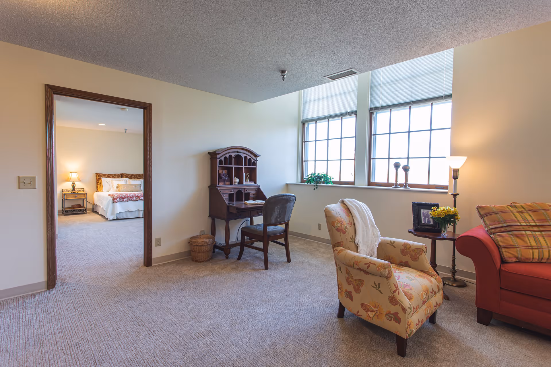 A cozy living room area with a floral armchair, a red sofa with a plaid cushion, a small round wooden side table with a framed photo and a vase of yellow flowers, and a floor lamp. There is a wooden desk with a chair against the wall near large windows with blinds. Through an open doorway, a bedroom with a bed, nightstand, and lamp is visible.