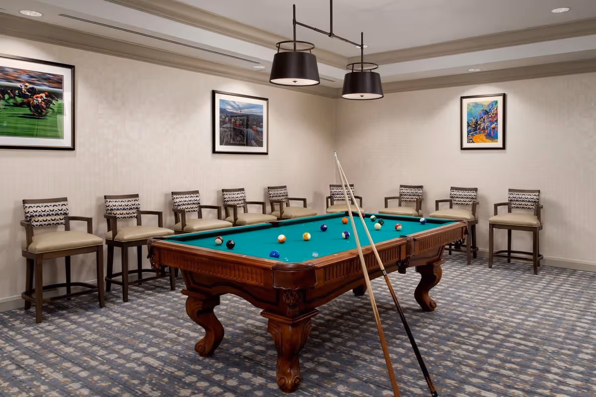A game room with a green felt pool table in the center, two pool cues resting on the table, and several billiard balls scattered on the table. The room has patterned carpet flooring, beige walls with three framed pictures, and a row of cushioned chairs with patterned backs lined up against the wall. Two black pendant lights hang from the ceiling above the pool table.