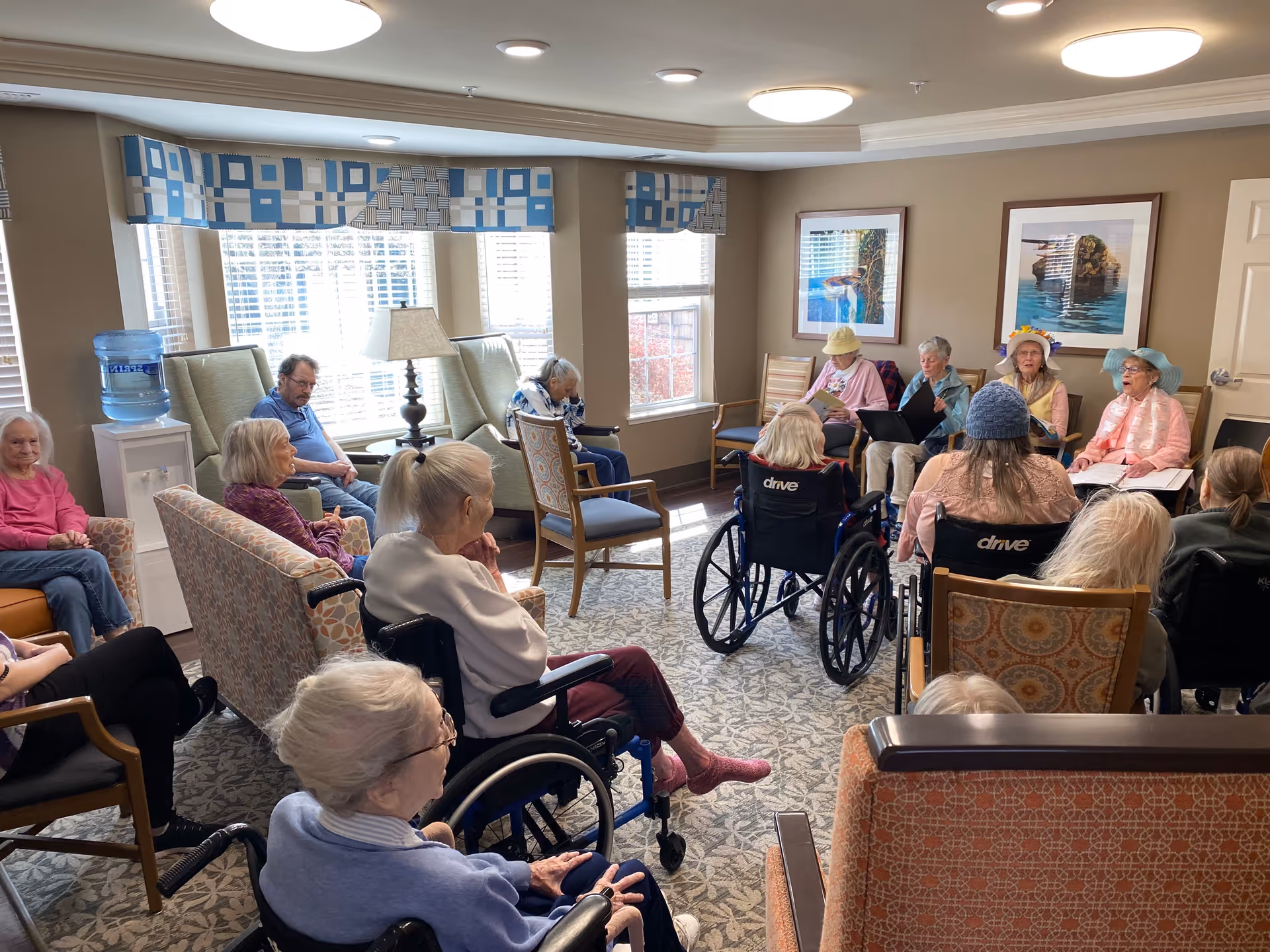 A group of elderly people seated in a well-lit common room with beige walls and patterned carpet. Some are in wheelchairs while others sit on armchairs arranged in a circle. Four women at the front appear to be leading an activity, wearing colorful hats and holding books or folders. Large windows with blue and white patterned valances let in natural light, and framed pictures hang on the walls.