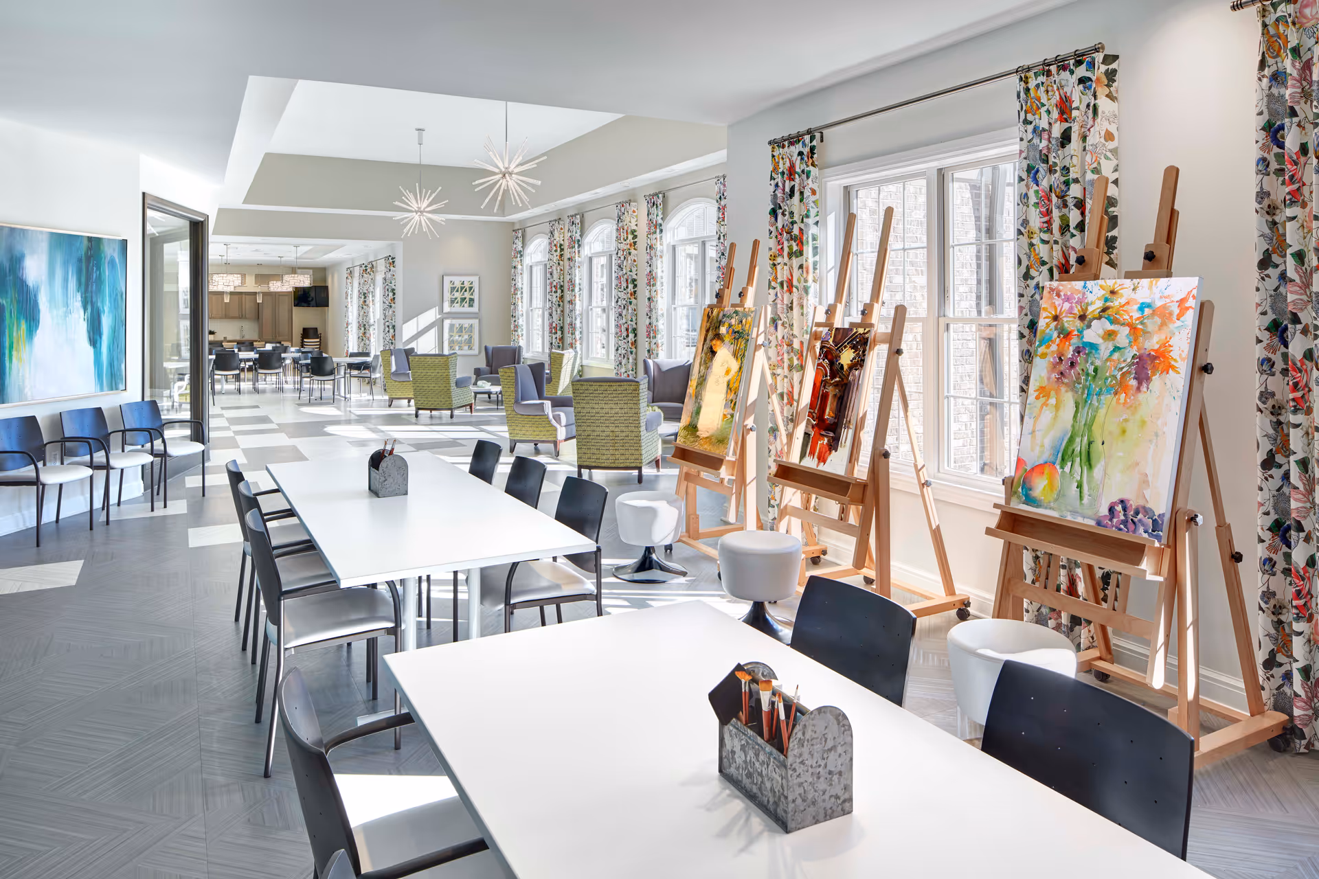 Bright and spacious art room with large windows, floral curtains, easels displaying colorful paintings, white tables with black chairs, and a seating area with green and purple armchairs in the background.