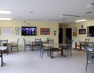 A dining room with several metal-framed tables and chairs arranged on a light-colored tiled floor. The walls are pale yellow and decorated with a 'Happy Birthday' sign, a bulletin board, and a mounted flat-screen TV displaying a group of people. There is a serving window on the right side and fluorescent ceiling lights.