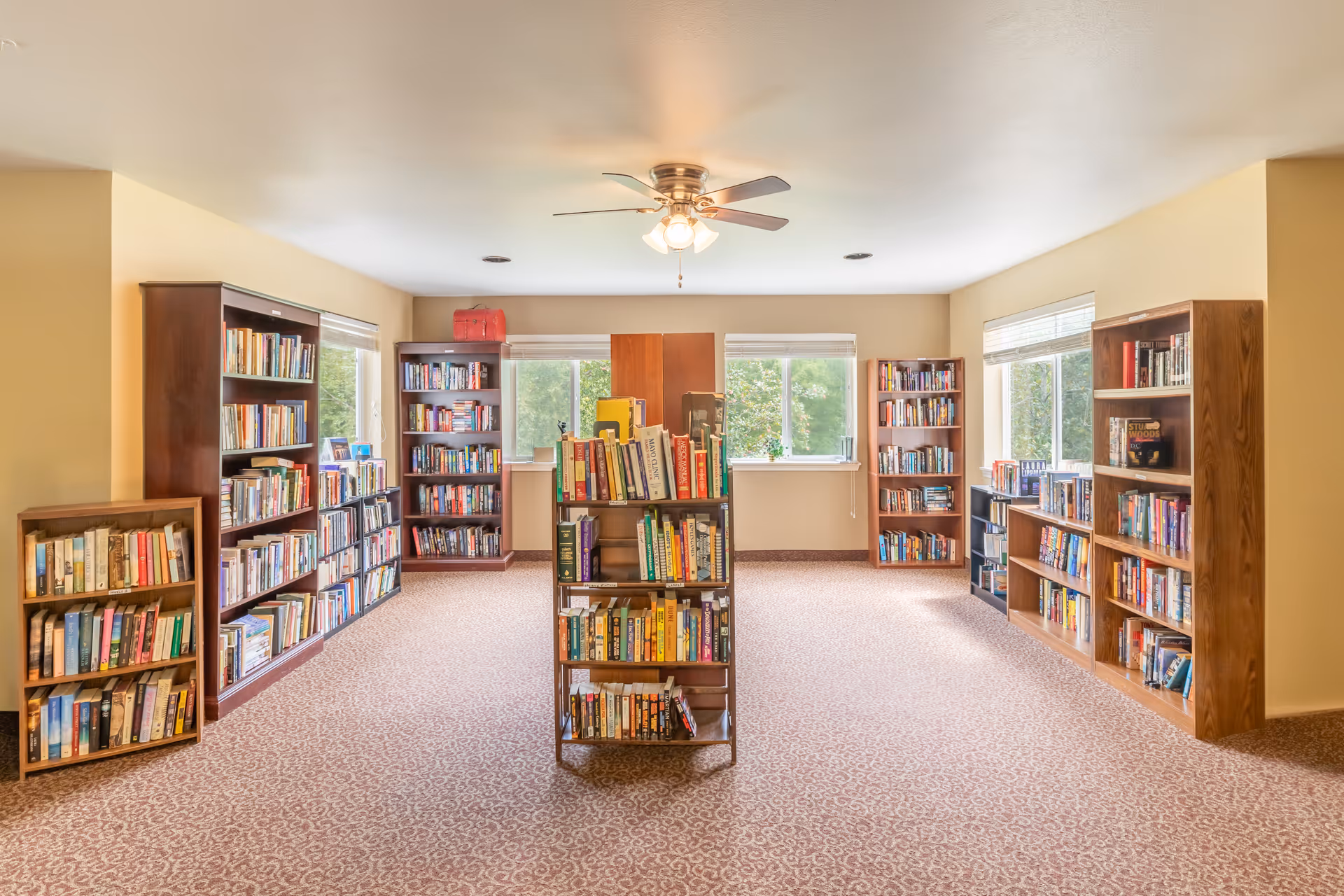 A bright and spacious room with multiple wooden bookshelves filled with books arranged along the walls and a central bookshelf. The room has beige walls, patterned carpet flooring, three large windows letting in natural light, and a ceiling fan with lights.