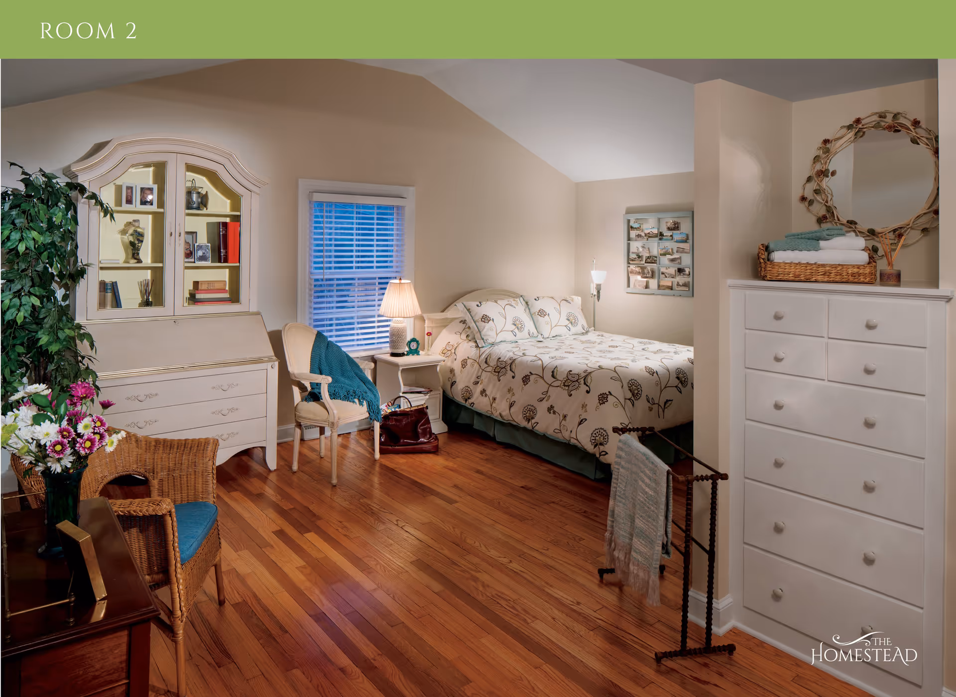 A cozy bedroom with hardwood floors featuring a bed with floral bedding, a white nightstand with a lamp, a white dresser with a decorative mirror above it, a wicker chair with a blue cushion, a white chair with a blue throw, a white cabinet with glass doors displaying books and photos, and a plant in the corner. There is a window with blinds and a photo collage on the wall.