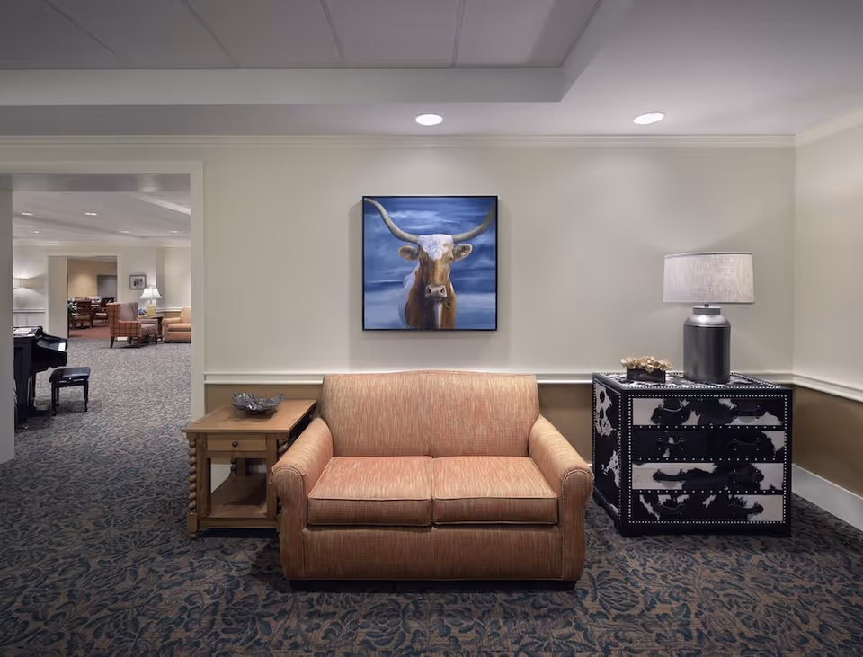A cozy seating area in a senior living facility featuring a peach-colored loveseat centered against a beige wall. Above the loveseat hangs a painting of a longhorn cow with a blue sky background. To the left of the loveseat is a wooden side table with a decorative bowl, and to the right is a black and white patterned chest with a lamp and small decorative items on top. The carpet has a floral pattern, and an open doorway reveals more seating and a piano in the adjacent room.