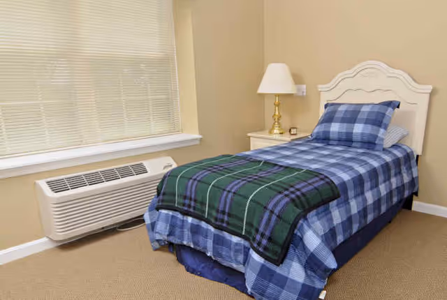 A single bed with a blue and green plaid blanket and matching pillow in a beige room. Next to the bed is a small nightstand with a brass lamp and a small clock. A window with closed blinds is above a wall-mounted air conditioning unit.