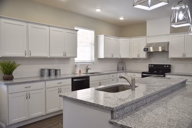 Bright modern kitchen with white cabinets, a granite island with sink, stainless steel appliances, and pendant lights.