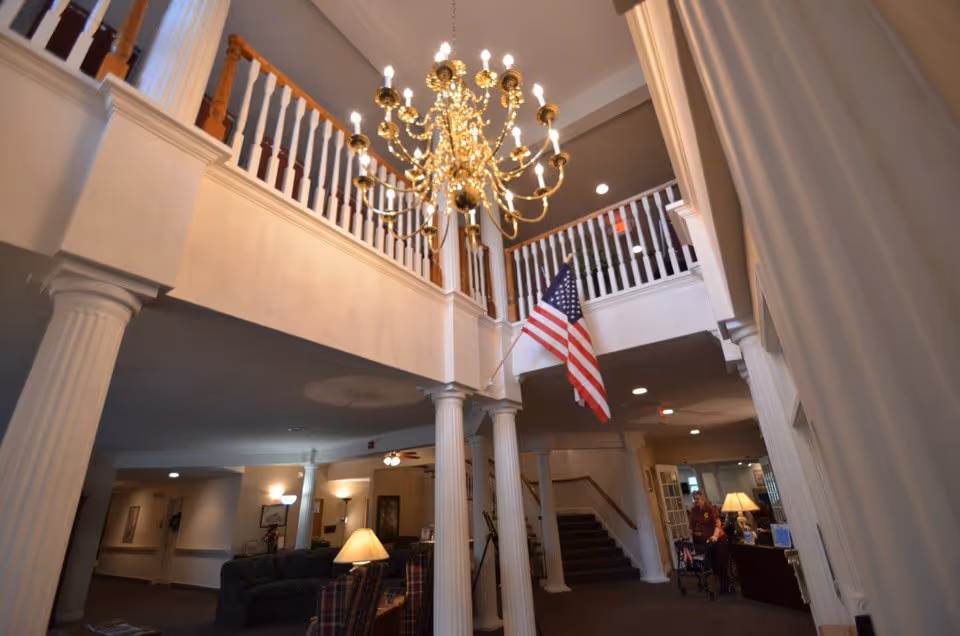 Interior lobby with a large chandelier, white columns, second-floor balcony, an American flag, and seating area.