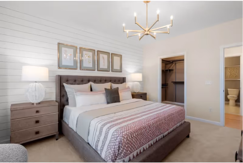 A neatly made bed with a tufted headboard in a bedroom featuring two bedside tables with lamps, four framed floral artworks above the bed, a modern chandelier, an open walk-in closet, and a bathroom visible through an open door.