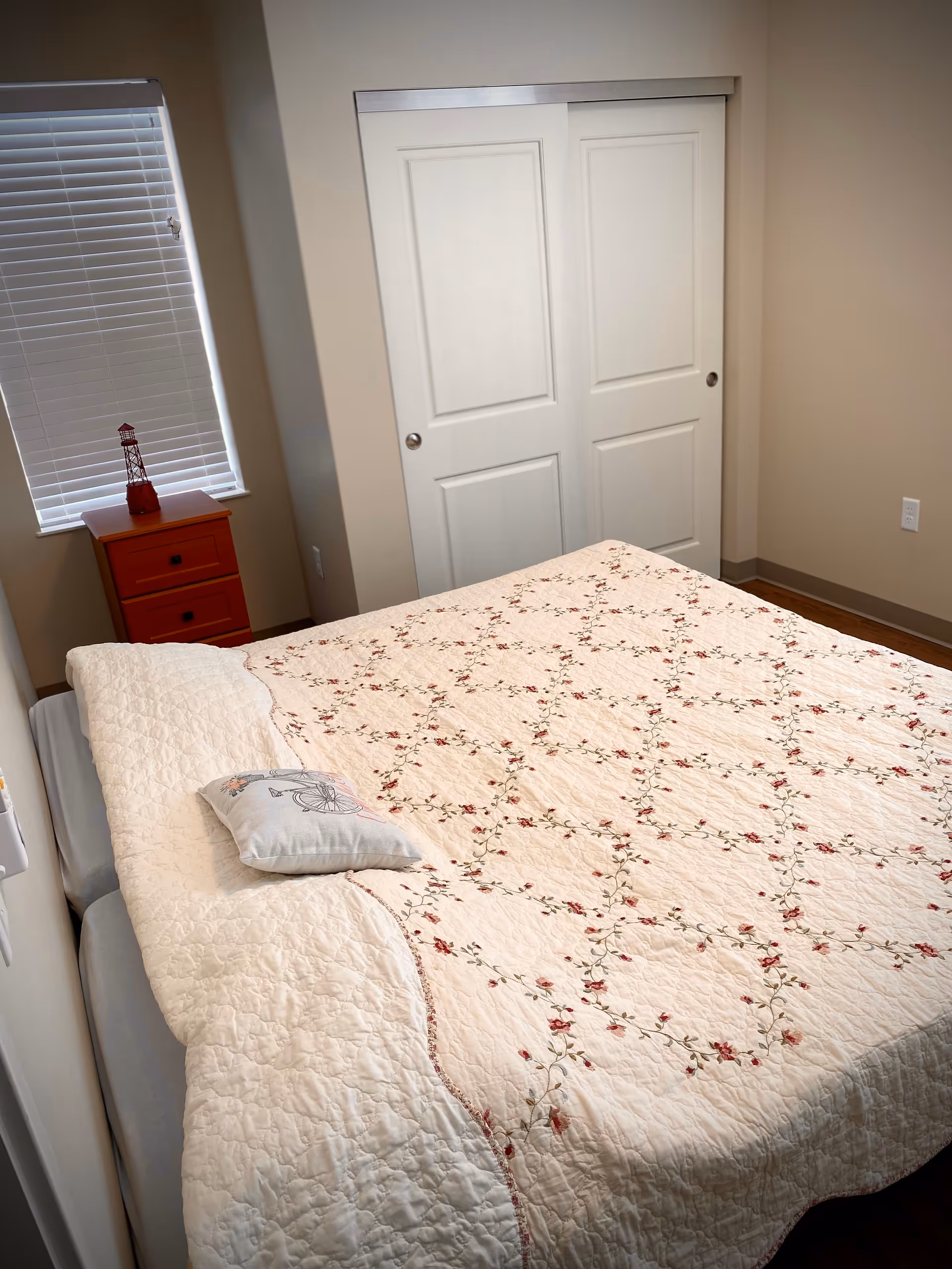 A bedroom with a bed covered in a cream-colored quilt with a floral pattern and a small decorative pillow. There is a wooden nightstand with two drawers next to a window with closed blinds. The room has beige walls and a white sliding closet door.
