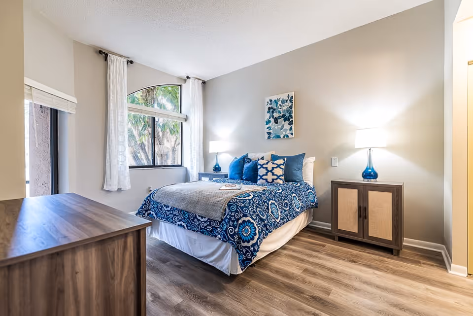 A bright and cozy bedroom with a bed dressed in blue and white patterned bedding and multiple pillows. The room features two bedside tables with matching blue lamps, a wooden dresser, light-colored walls, wood flooring, and a large window with white curtains allowing natural light to enter.