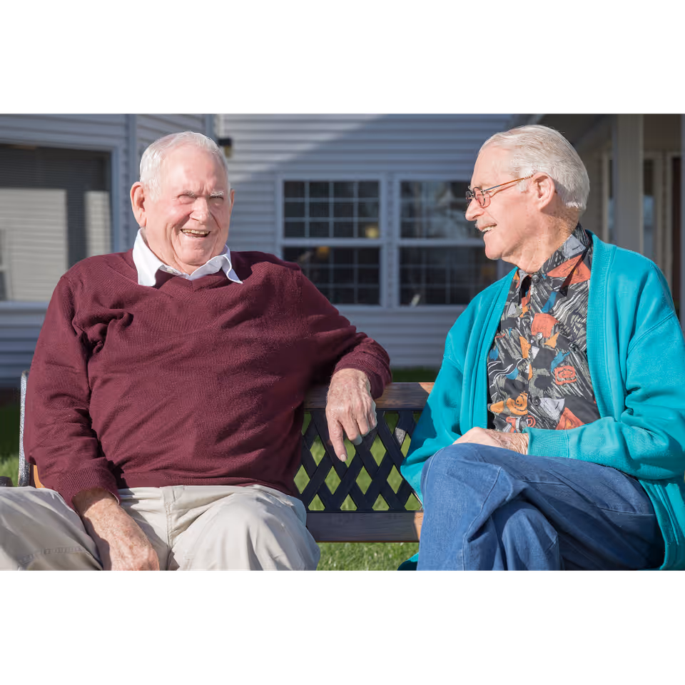 Two elderly men sitting on a bench outside a building, smiling and engaging in conversation. One man is wearing a maroon sweater and light-colored pants, while the other is wearing a teal cardigan over a patterned shirt and blue pants. The background shows windows and siding of the building.