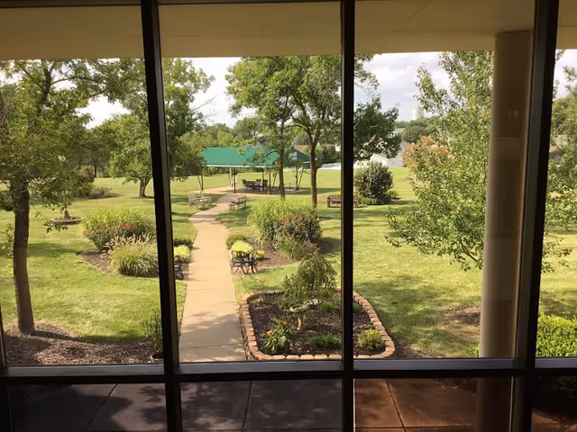 View through large window panes showing a landscaped outdoor area with a paved walkway, benches, trees, and a green canopy-covered seating area in the distance.