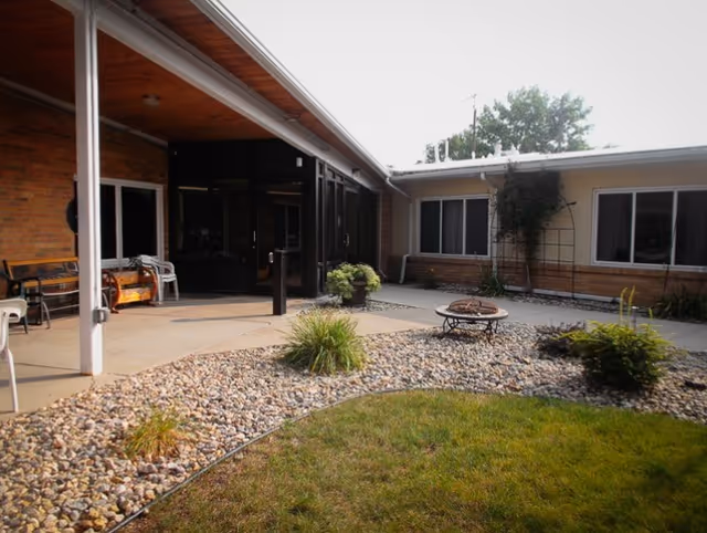 Outdoor courtyard area of a senior living facility with a covered patio, chairs, benches, small plants, a fire pit, and windows of the building surrounding the courtyard.