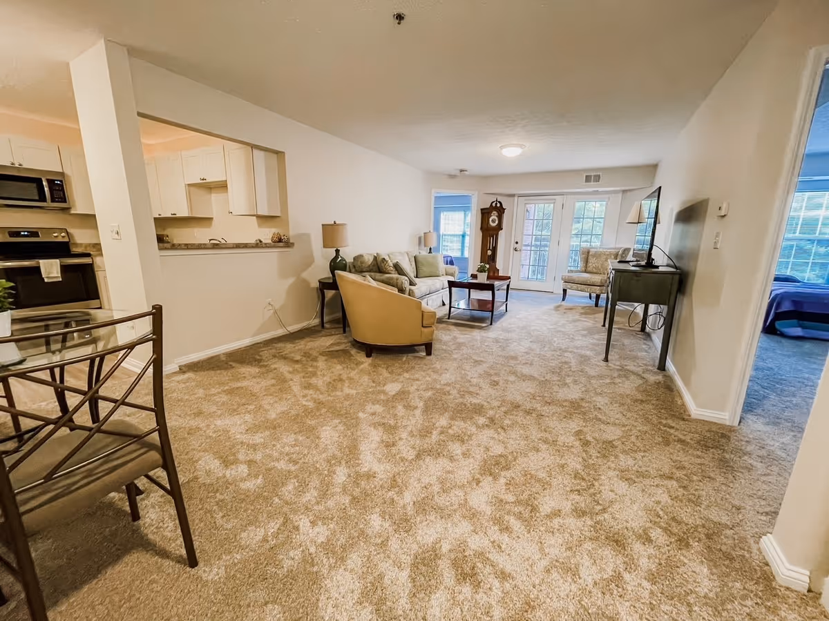 A spacious living room with beige carpet, a sofa, armchair, coffee table, and a TV on a stand. The room has a grandfather clock near glass doors leading outside. To the left is a kitchen area with white cabinets and stainless steel appliances. A dining chair is partially visible in the foreground. A bedroom with a bed and window is visible through an open doorway on the right.