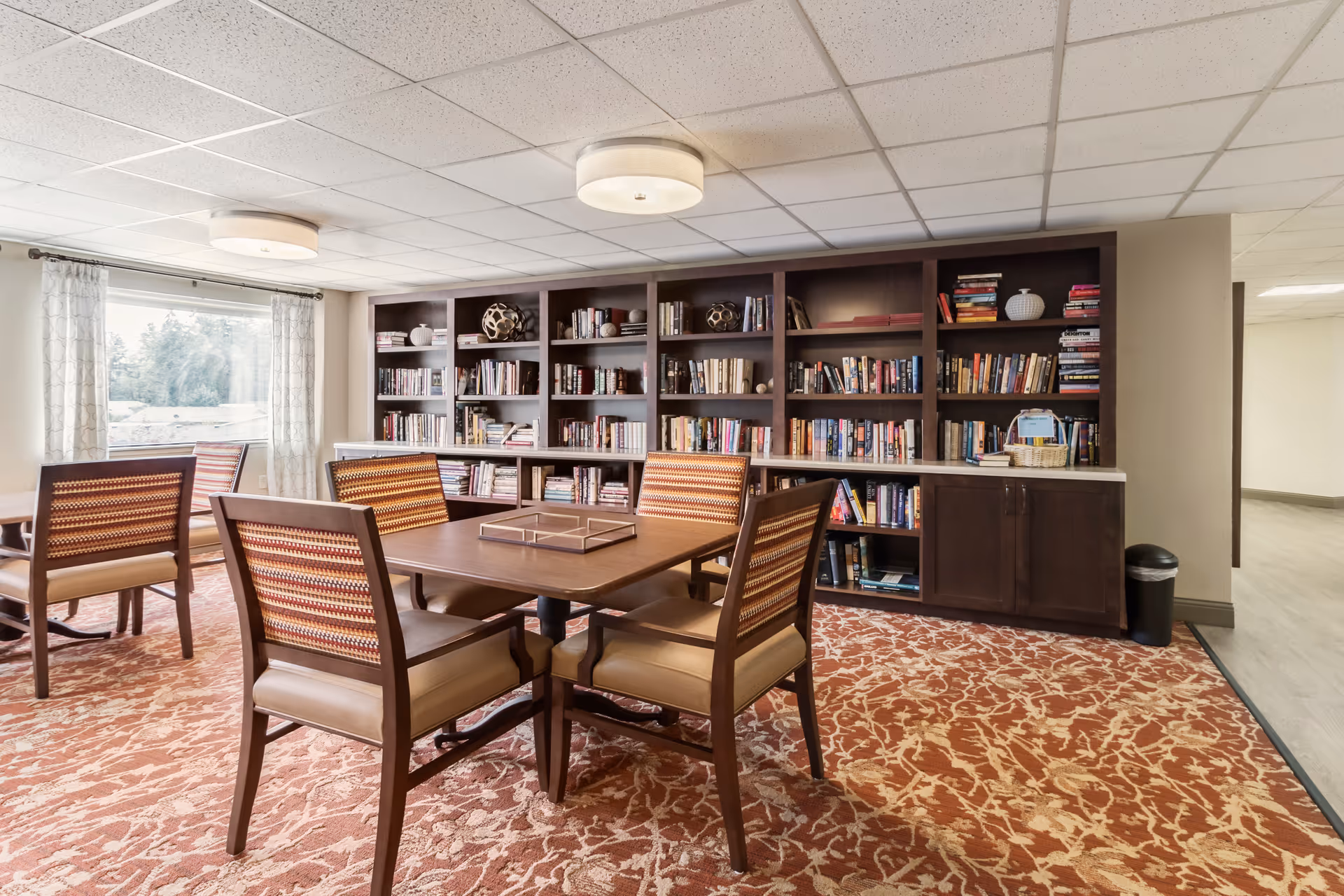A bright common room with a wooden table and chairs facing built-in bookshelves filled with books.