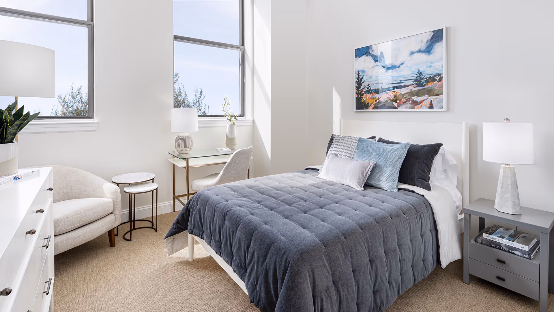 A bright and cozy bedroom with a neatly made bed featuring a dark gray quilt and multiple pillows. The room has two large windows letting in natural light, a white armchair, a small set of nesting tables, a glass desk with a chair, and a gray nightstand with a lamp and books. A colorful landscape painting hangs on the wall above the bed.
