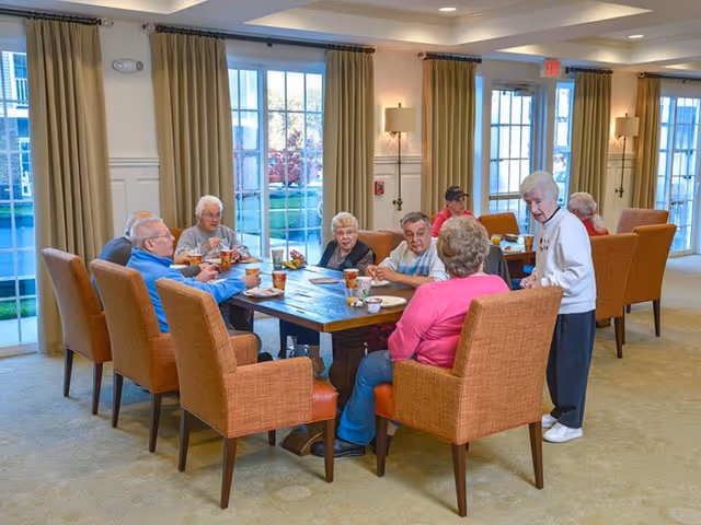 A group of elderly residents sitting around a long dining table in a bright communal room with large windows and orange upholstered chairs.