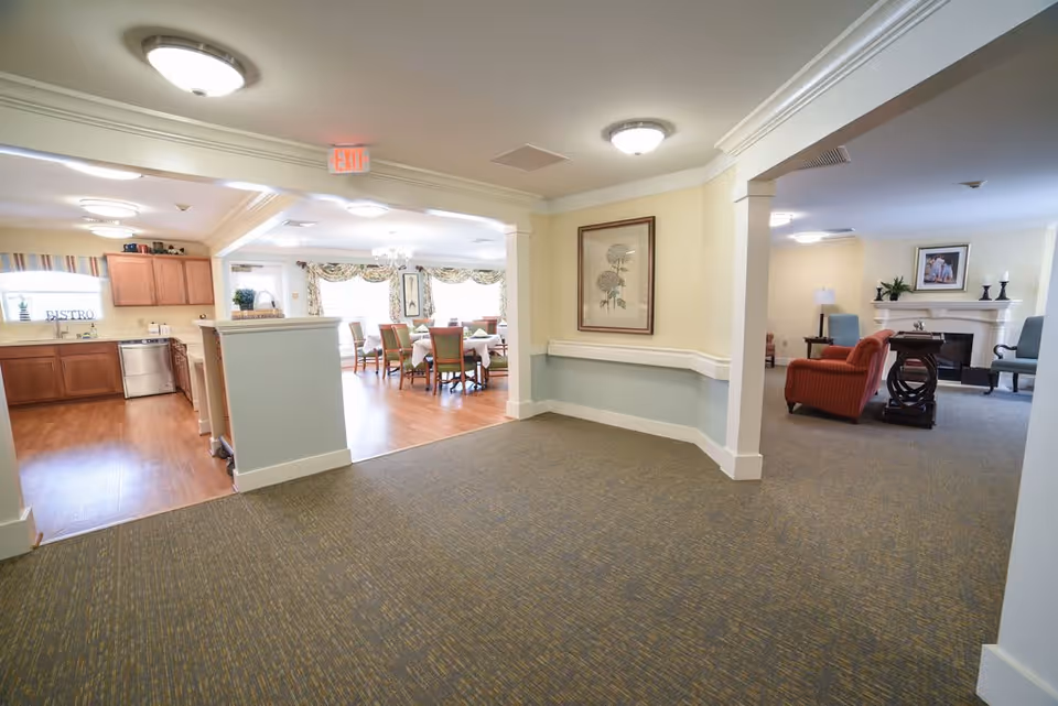 Interior view of a senior living facility showing a hallway leading to a dining area with tables and chairs, a kitchen area with wooden cabinets and a dishwasher on the left, and a living room area with armchairs and a fireplace on the right. The walls are painted light yellow and light blue, and the floor is carpeted in the hallway with wood flooring in the kitchen and dining areas.