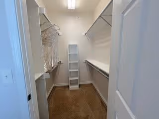 A walk-in closet with beige carpet flooring, white walls, and white shelving units on both sides. There is a tall, narrow white shelving unit at the far end of the closet. The closet is well-lit with a ceiling light fixture.