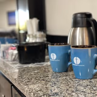 Close-up view of a granite countertop with two blue coffee mugs labeled 'Presbyterian Horizons', a stainless steel thermal coffee carafe, and a black container holding sugar packets and stirrers in the background.