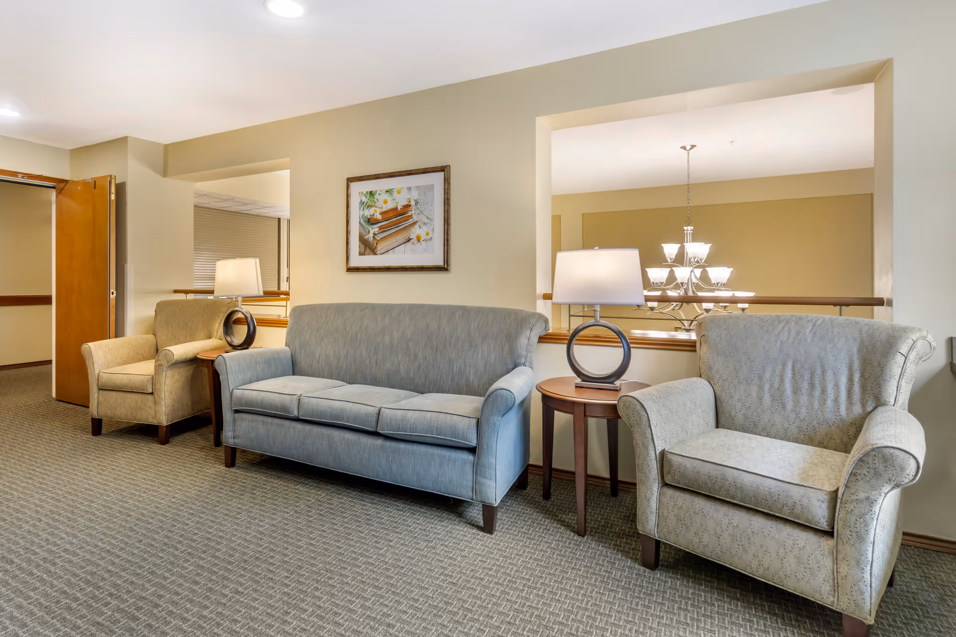 A seating area in a senior living facility featuring a blue upholstered sofa, two beige armchairs, and two round wooden side tables with modern lamps. The walls are painted light beige, and there is a framed picture of books and flowers hanging on the wall. A chandelier is visible through a large interior window opening.