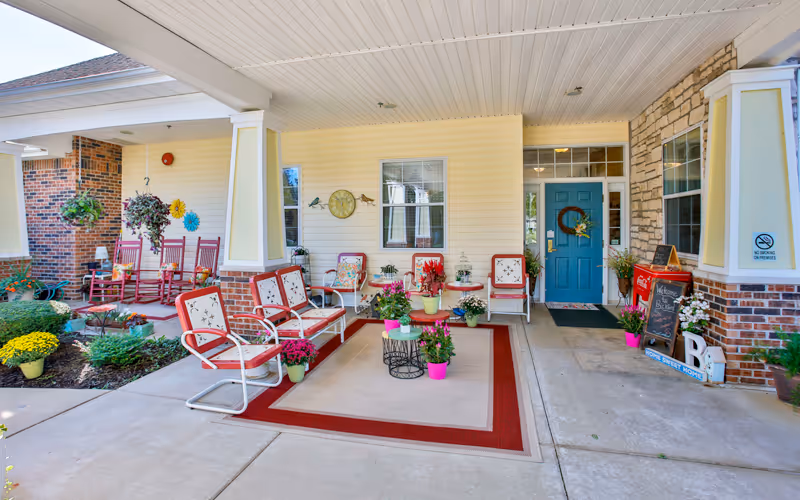 Covered outdoor patio area at 1019 Belle's Place of Crawfordsville with several cushioned chairs and rocking chairs arranged around a large area rug. The space is decorated with multiple potted plants and flowers. A blue door with a wreath and a welcome sign is visible, along with a no smoking sign on the wall.