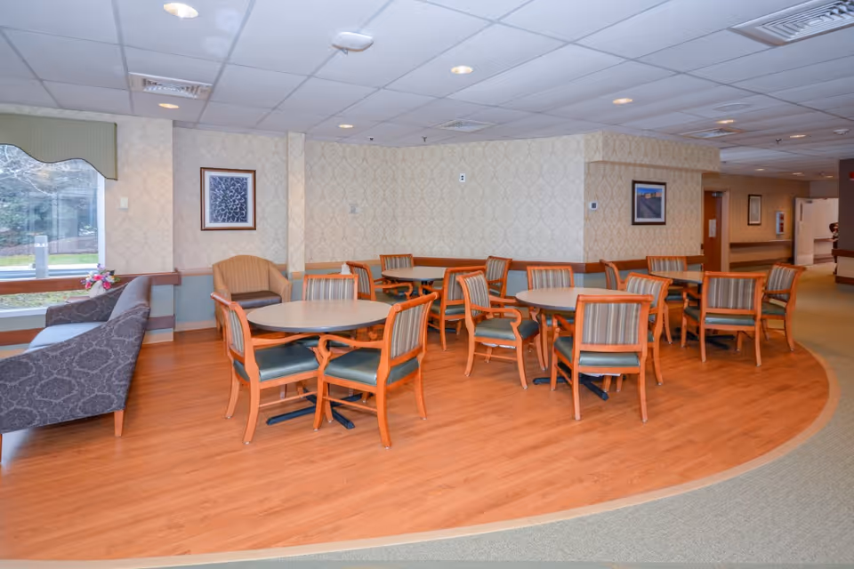 A common area in a senior living facility with several round tables surrounded by wooden chairs with green cushions. There is a patterned sofa and an armchair near a large window with a view of the outdoors. The walls are decorated with framed artwork and wallpaper with a subtle pattern. The floor is a combination of wood and carpet.
