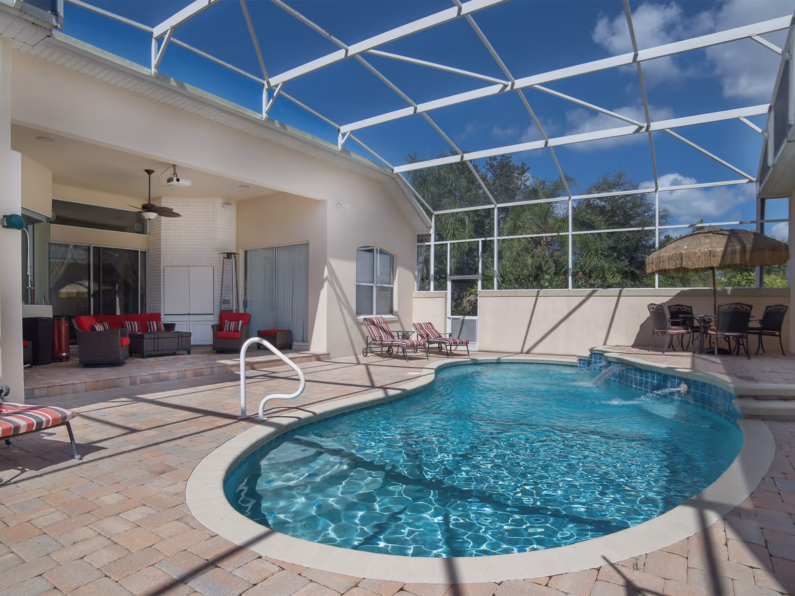 A screened-in outdoor pool area with clear blue water, surrounded by a brick patio. There are several lounge chairs with red and white striped cushions, a seating area with red cushioned wicker furniture, and a table with chairs under a thatched umbrella. The sky is blue with some clouds visible through the screen enclosure.