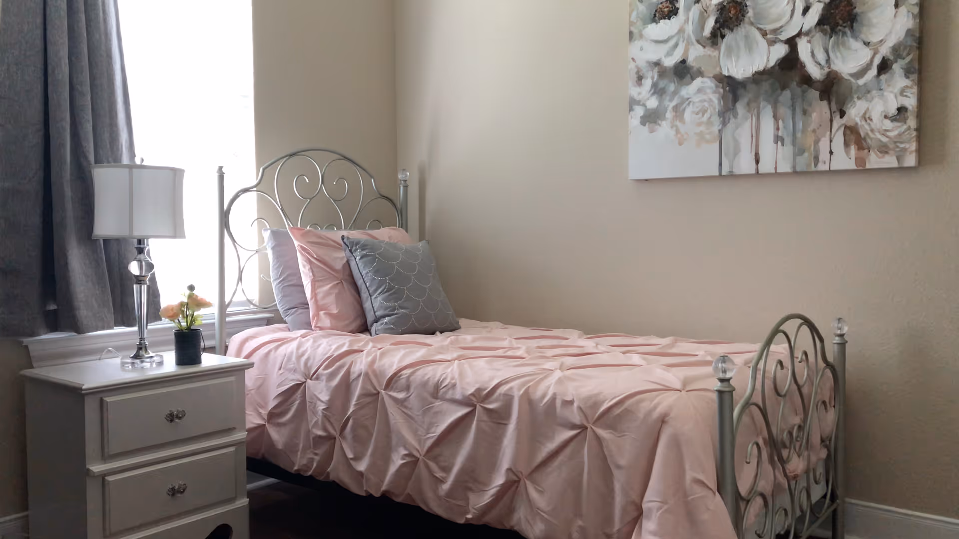 Small bedroom with a single metal-framed bed dressed in pink bedding, a white nightstand with a lamp and flowers, and floral wall art above the bed.