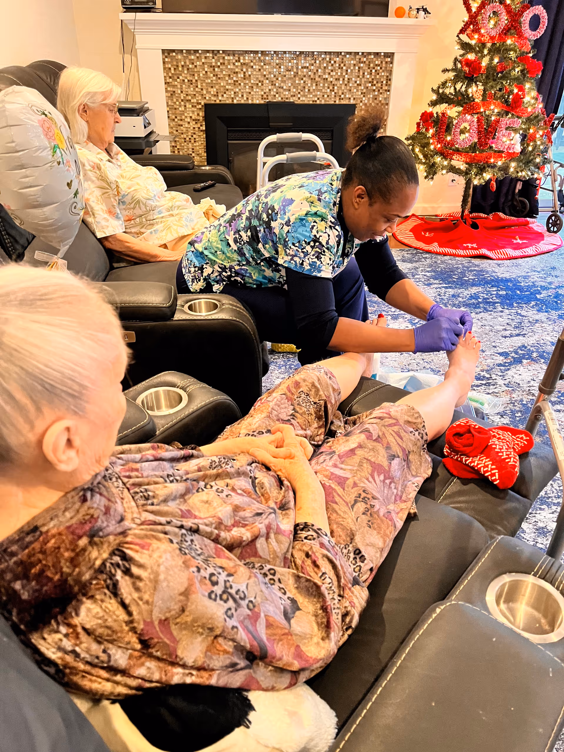 A caregiver wearing gloves is providing foot care to an elderly woman seated in a reclining chair in a cozy living room. Another elderly woman is seated nearby on a similar chair. A decorated Christmas tree with red and white ornaments and a 'LOVE' sign is visible in the background near a fireplace.