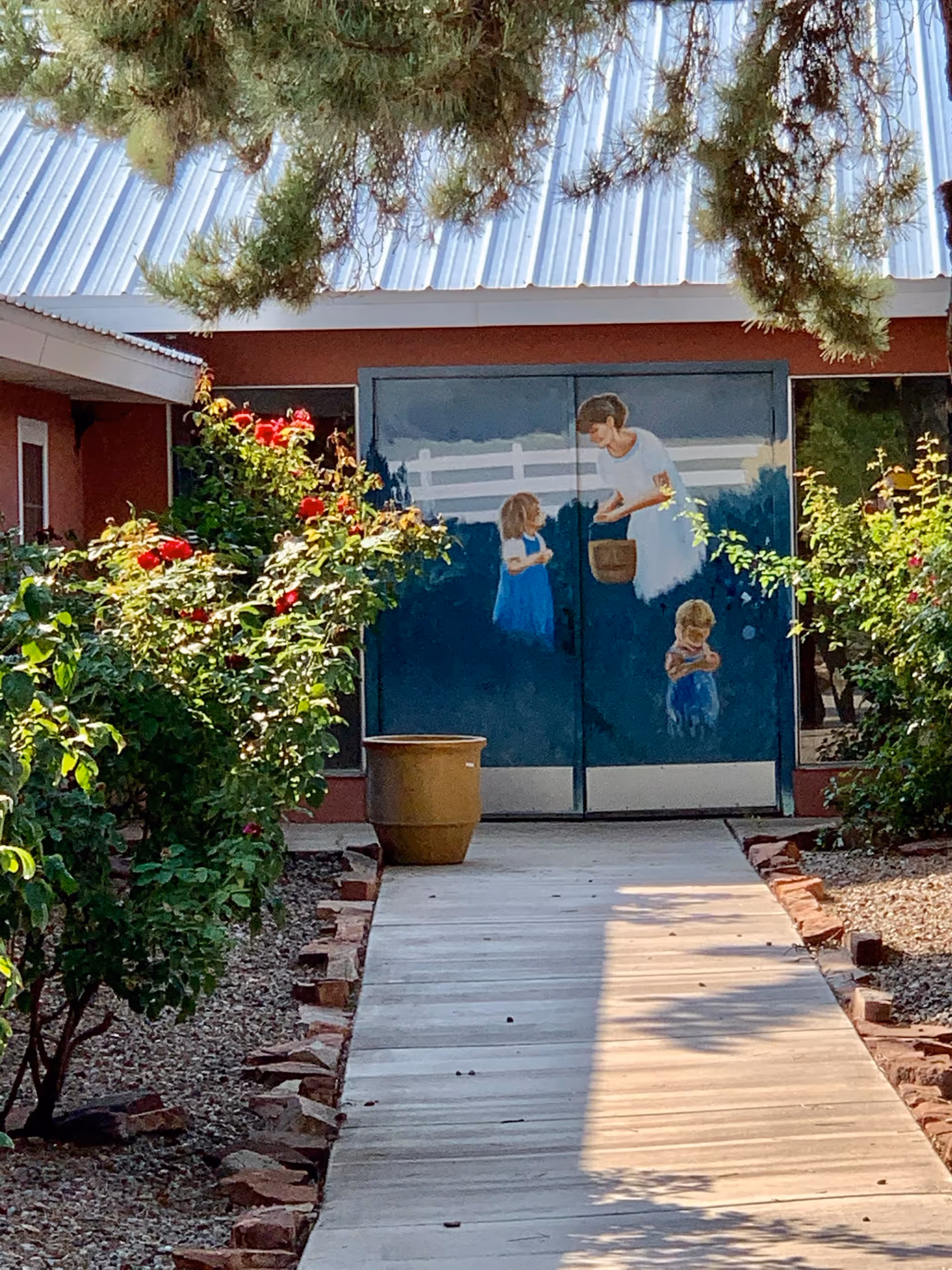 A garden pathway leading to a building entrance with a mural on the doors depicting a woman and two children. The pathway is bordered by rose bushes with red flowers and a large planter is placed near the door. The building has a metal roof and is partially shaded by tree branches.