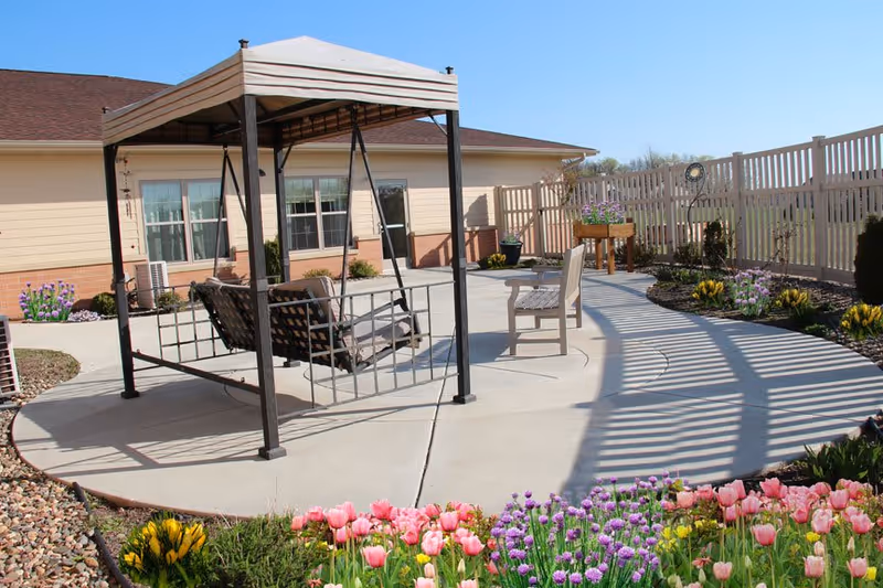 Outdoor patio area with a metal-framed swing bench under a canopy, a wooden bench, and a curved concrete walkway surrounded by flower beds with colorful tulips and other flowers. A beige building with windows and a door is in the background, along with a white fence enclosing the area.