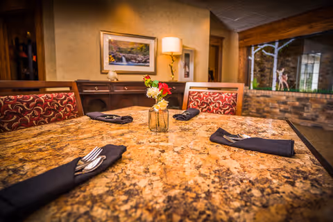 A dining table with a polished granite surface set with black napkins holding silverware, and a small vase with colorful flowers in the center. The background shows a wooden sideboard, a lamp, a framed picture on the wall, and a window with a view of a stone wall and outdoor greenery.