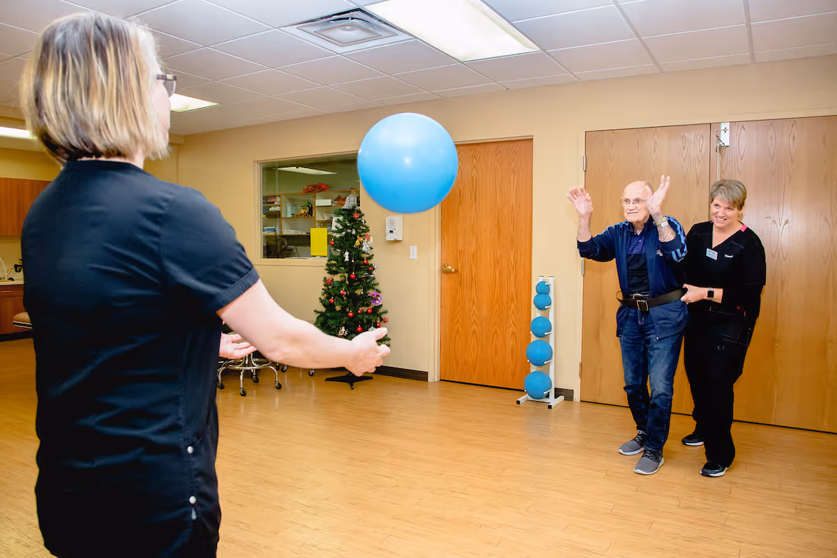 An elderly man is assisted by a caregiver as he plays with a large blue balloon in a spacious room with wooden floors and beige walls. Another person is throwing the balloon towards them. A small decorated Christmas tree and a rack with blue exercise balls are visible in the background.