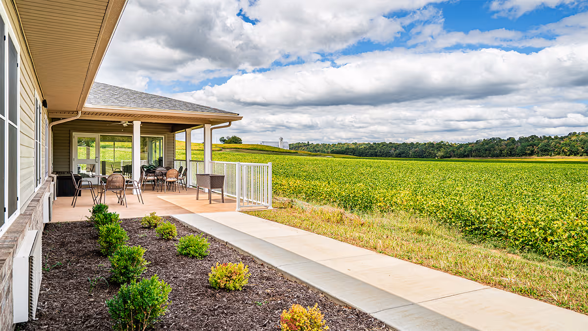 Covered patio area with several tables and chairs next to a building, overlooking a large green field under a partly cloudy sky.