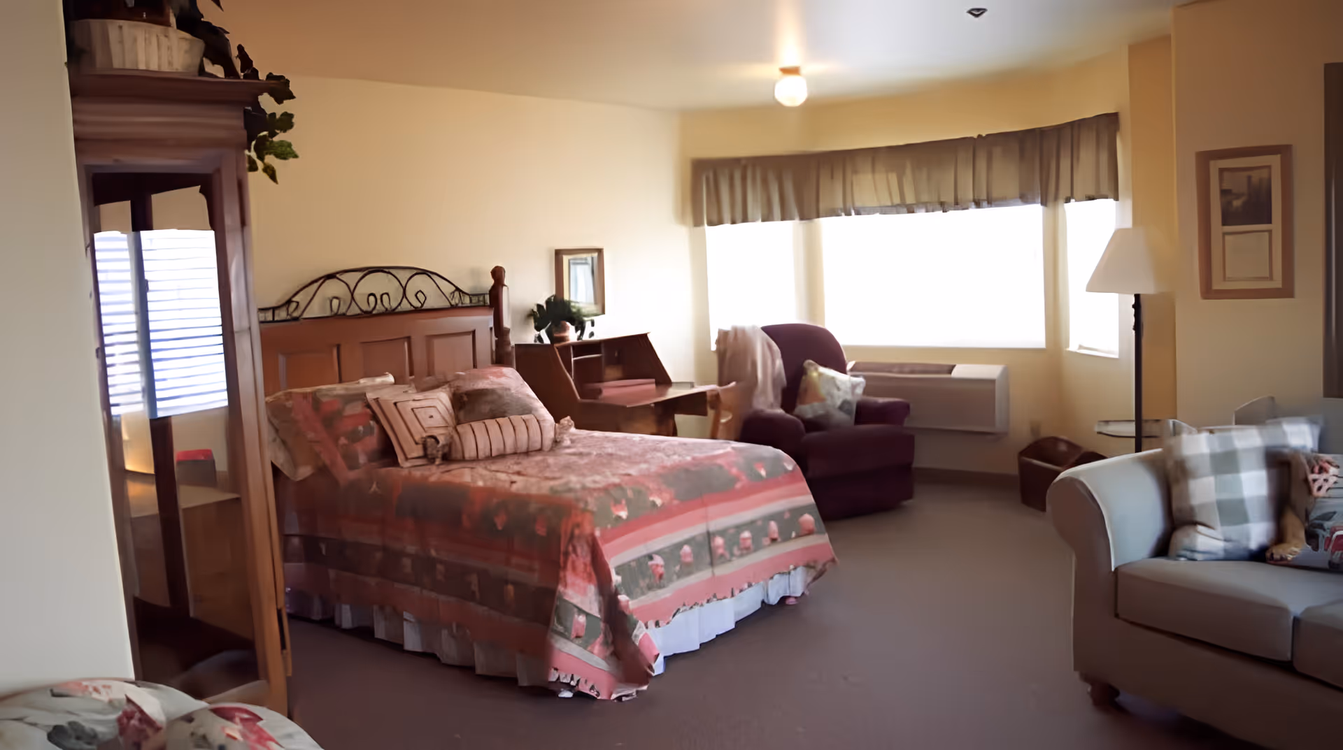 A cozy bedroom in an assisted living facility featuring a wooden bed with a patterned bedspread, a purple armchair with a throw blanket, a small wooden desk, a floor lamp, a sofa with plaid pillows, and large windows with curtains letting in natural light.