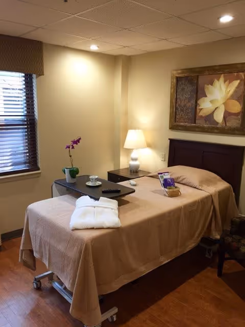 A single bed in a cozy room with beige walls and wooden flooring. The bed is covered with a light brown blanket and has a white folded towel on it. A small table on wheels is placed over the bed, holding a potted orchid, a cup and saucer, and a remote control. Next to the bed is a nightstand with a white lamp and a framed floral painting hanging above the headboard. A window with wooden blinds is on the left side of the room.